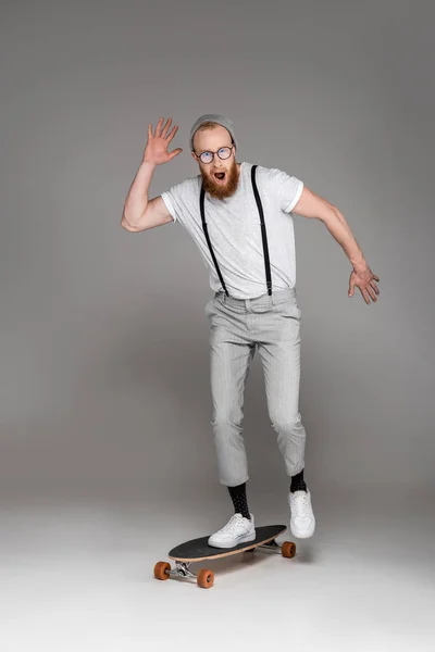 Scared bearded man with open mouth standing on longboard and looking at camera on grey — Stock Photo