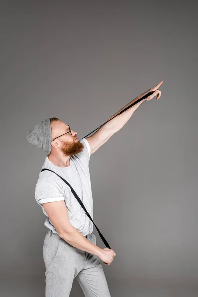 Hombre barbudo con estilo sosteniendo tirantes y apuntando lejos aislado en gris - foto de stock