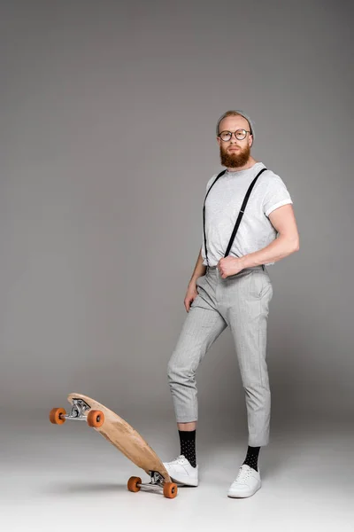 Стильный бородатый мужчина в подтяжках, стоящий со скейтбордом и смотрящий в камеру на сером — стоковое фото