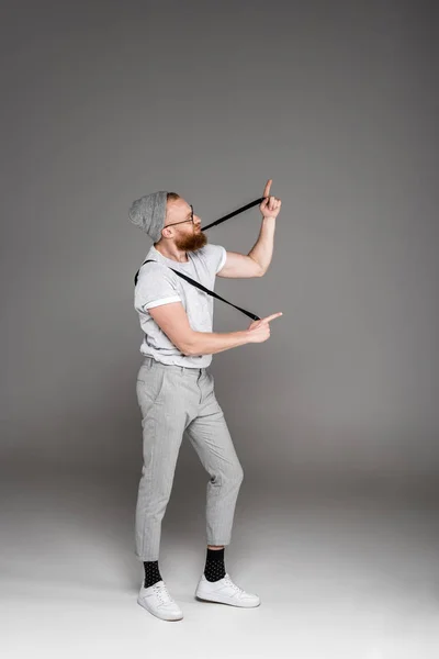 Homem barbudo elegante jogando com suspensórios e apontando com os dedos no cinza — Fotografia de Stock