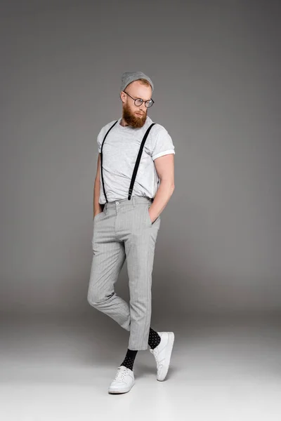 Handsome bearded man in suspenders and spectacles standing with hands in pockets and looking down on grey — Stock Photo