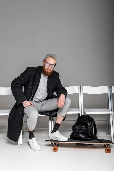 Elegante uomo barbuto in cappotto seduto su sedie con longboard e zaino in pelle su grigio — Foto stock