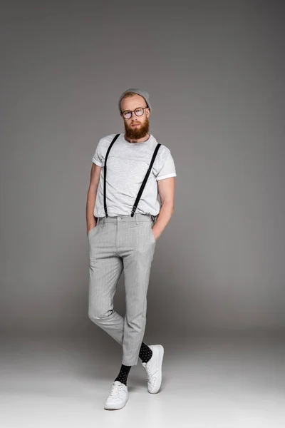 Elegante uomo barbuto in cappello e occhiali in piedi con le mani in tasca e guardando la fotocamera su grigio — Foto stock