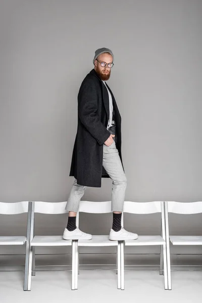 Élégant homme barbu en chapeau, lunettes et manteau marchant sur des chaises et regardant la caméra sur gris — Photo de stock