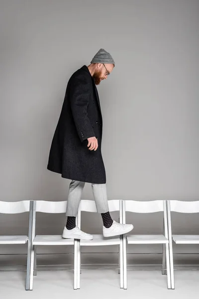 Vue latérale de l'homme barbu élégant en chapeau, lunettes et manteau marchant sur des chaises sur gris — Photo de stock