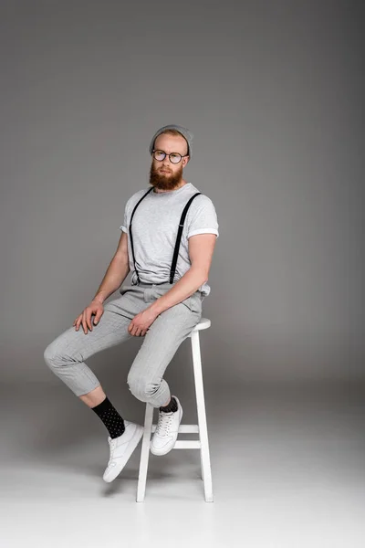 Stylish bearded hipster in eyeglasees sitting on stool and looking at camera on grey — Stock Photo