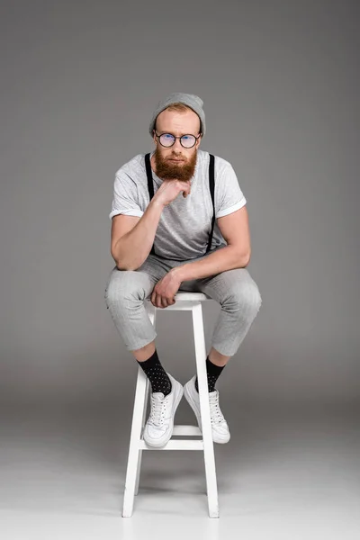 Stylish bearded man in eyeglasees sitting on stool and looking at camera on grey — Stock Photo