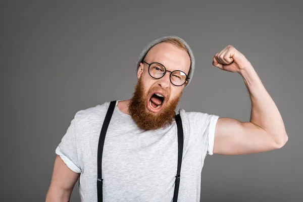 Excited bearded man showing biceps and screaming at camera isolated on grey — Stock Photo