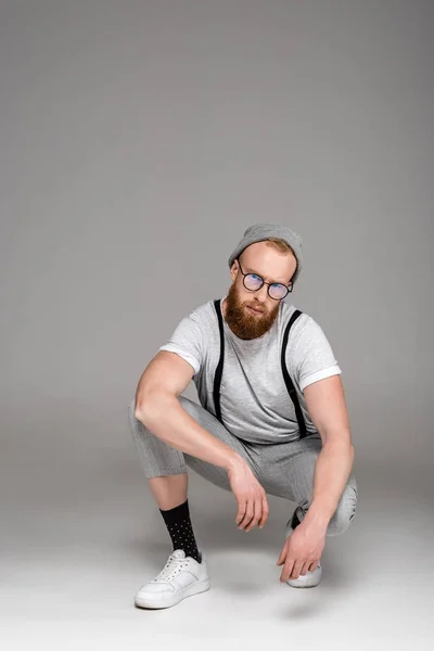 Стильный бородатый мужчина в шляпе и очках, приседающий и смотрящий на камеру на сером — стоковое фото