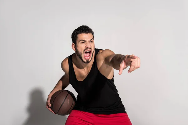 Агрессивный молодой спортсмен, держащий баскетбольный мяч, указывающий пальцем и кричащий на белый — стоковое фото
