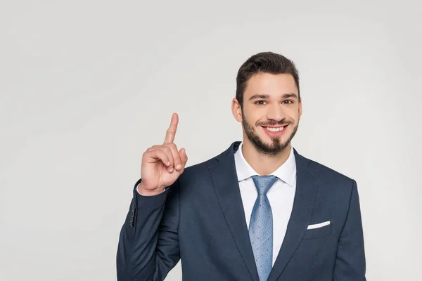 Lächelnder junger Geschäftsmann zeigt mit dem Finger nach oben und blickt isoliert in die Kamera auf grau — Stockfoto