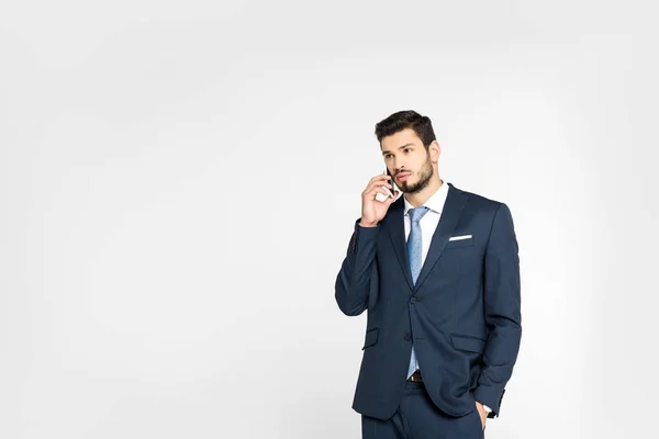 Joven y elegante hombre de negocios de pie con la mano en el bolsillo y hablando en el teléfono inteligente mientras mira hacia otro lado aislado en gris - foto de stock