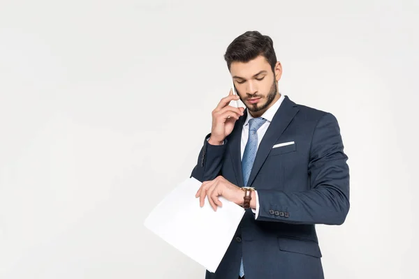 Junger Geschäftsmann hält Papiere in der Hand und telefoniert mit dem Smartphone, während er die Armbanduhr isoliert auf grau überprüft — Stockfoto