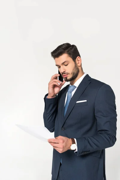 Молодий бізнесмен говорить на смартфоні і дивиться на папір ізольовано на сірому — стокове фото