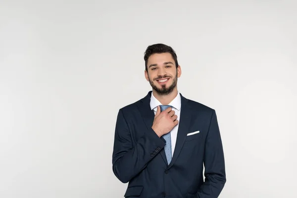 Handsome young businessman adjusting necktie and smiling at camera isolated on grey — Stock Photo