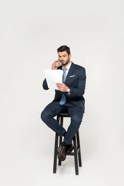 Serio joven hombre de negocios hablando en el teléfono inteligente y la lectura de papeles mientras está sentado en el taburete aislado en gris - foto de stock