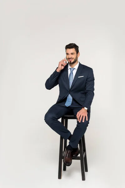 Smiling young businessman talking on smartphone while sitting on stool and looking away isolated on grey — Stock Photo