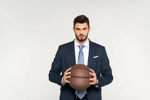 Serio joven empresario sosteniendo pelota de baloncesto y mirando a la cámara aislada en gris - foto de stock