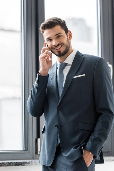 Молодой бизнесмен разговаривает на смартфоне и улыбается на камеру в офисе — стоковое фото
