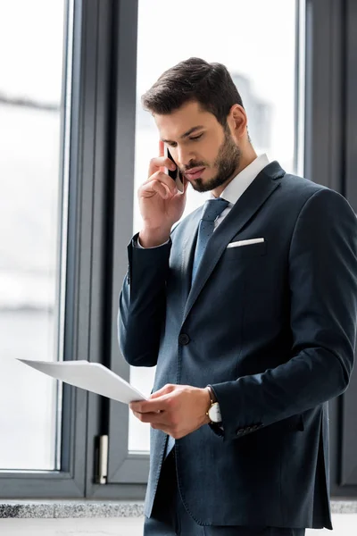 Fokussierter junger Geschäftsmann hält Papiere in der Hand und telefoniert im Büro mit dem Smartphone — Stockfoto