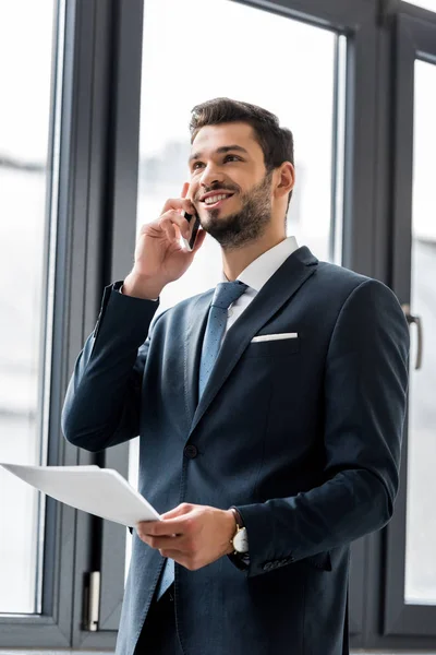 Sonriente joven empresario sosteniendo papeles y hablando en el teléfono inteligente en la oficina - foto de stock