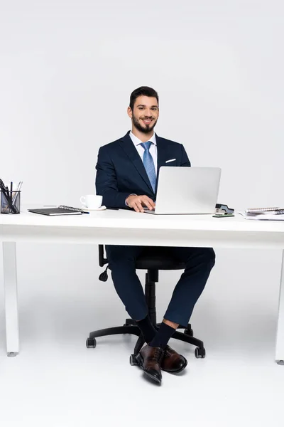 Handsome young businessman smiling at camera while using laptop at workplace — Stock Photo