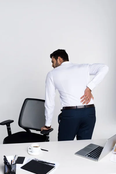 Молодий бізнесмен, який страждає від беккача, спираючись на стілець в офісі — стокове фото