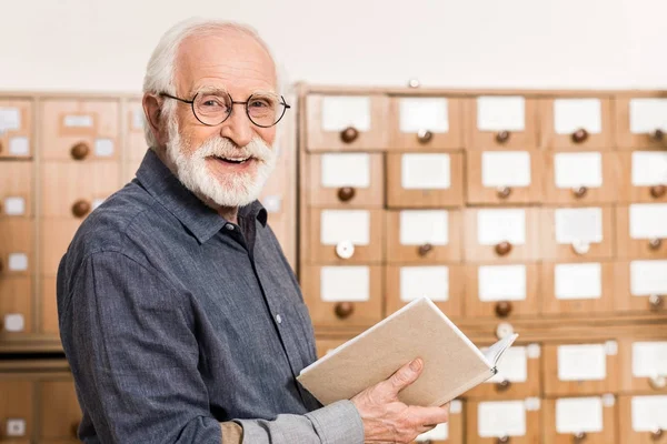 Sonriendo anciano archivista masculino sosteniendo libro y mirando a la cámara - foto de stock