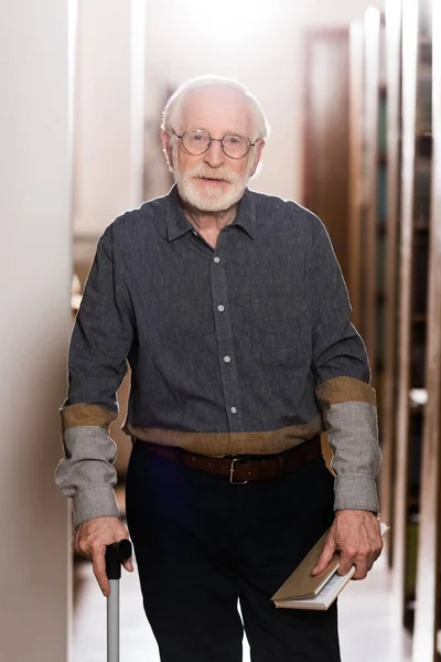 Grey hair librarian with walking stick and book looking at camera — Stock Photo