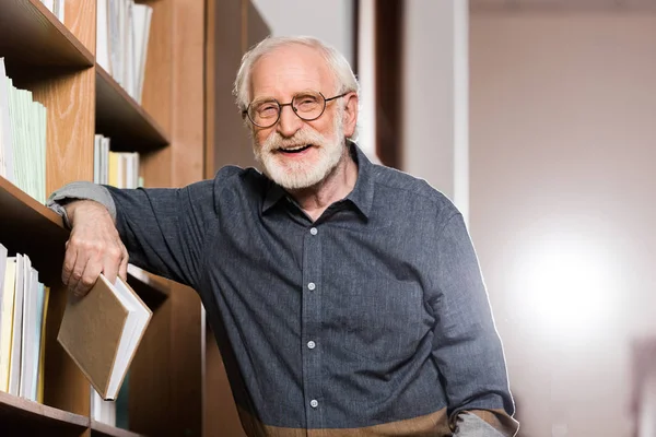 Graue Haare Bibliothekarin hält Buch in der Hand und lehnt an Regal — Stockfoto
