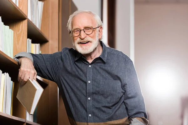 Bibliotecario capelli grigi che tiene libro e appoggiato sullo scaffale — Foto stock