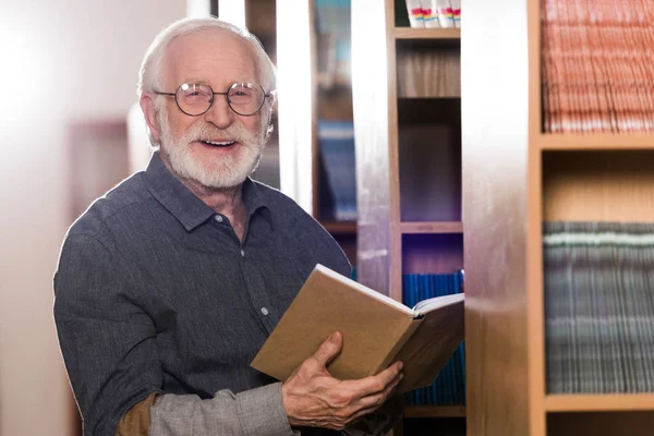 Souriant gris bibliothécaire cheveux tenant le livre et regardant la caméra — Photo de stock