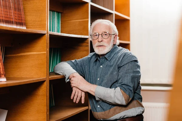 Graue Haare Bibliothekarin lehnt an Regalen und blickt in die Kamera — Stockfoto