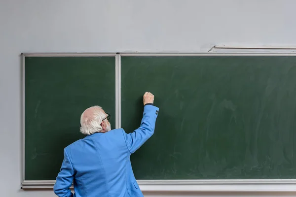 Vista posterior del profesor senior escribiendo algo en pizarra con tiza - foto de stock