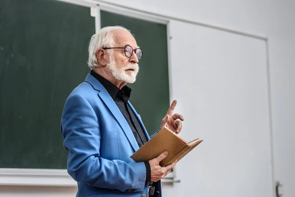 Profesor senior sosteniendo libro y mostrando un dedo hacia arriba - foto de stock