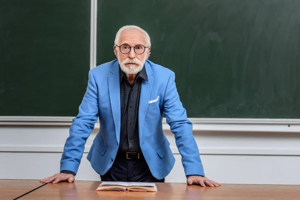 Profesor senior apoyado en la mesa y mirando hacia otro lado - foto de stock