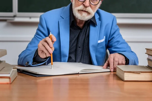 Обрезанный образ вдумчивого старшего преподавателя, сидящего за столом с карандашом и блокнотом — стоковое фото