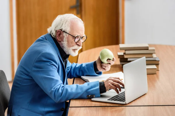 Професор сірого волосся тримає яблуко і використовує ноутбук — Stock Photo