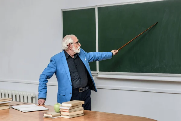 Professeur de cheveux gris pointant sur quelque chose au tableau noir — Photo de stock