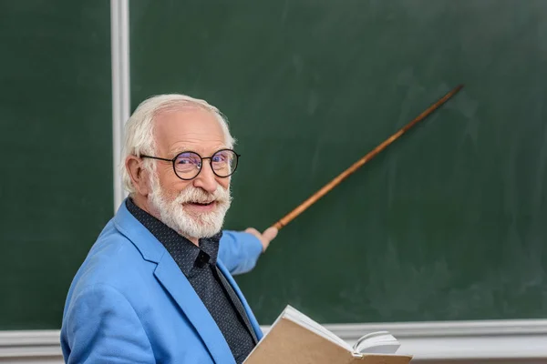 Grey hair professor holding book and pointing on something at blackboard — Stock Photo