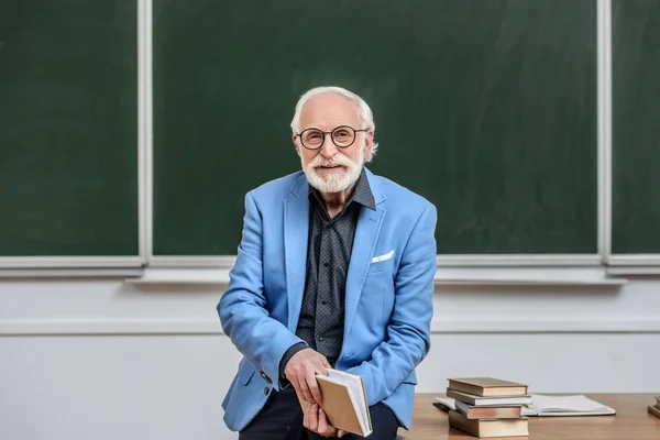 Professor de cabelos grisalhos sentado na mesa na sala de aula e livro segurando — Fotografia de Stock