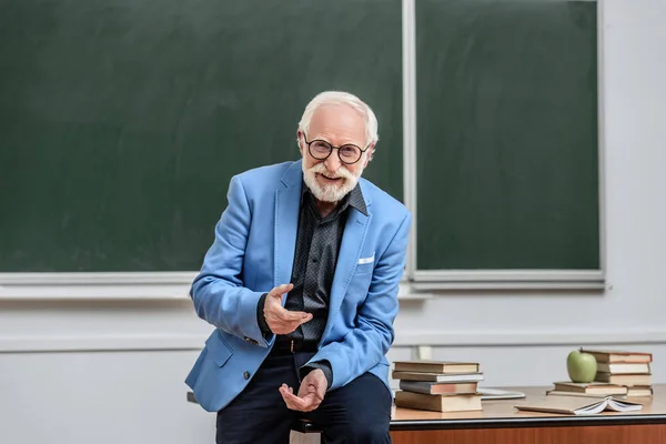 Smiling grey hair professor sitting on table in lecture room — Stock Photo