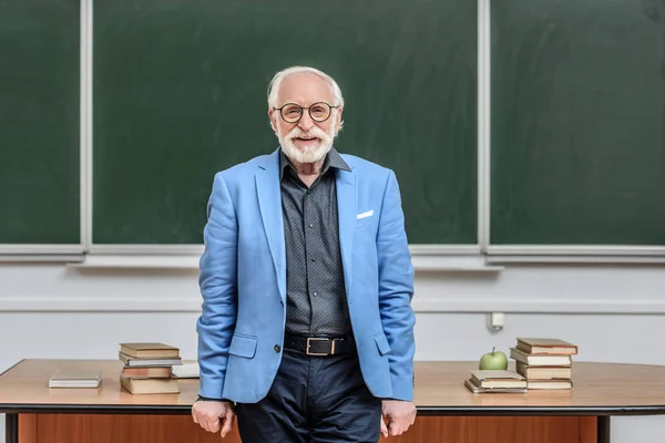 Grinsender Professor mit grauen Haaren steht im Hörsaal und blickt in die Kamera — Stockfoto