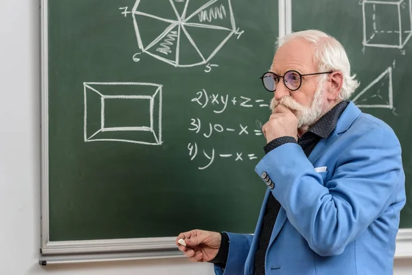 Nachdenkliche graue Haare Professor steht an der Tafel mit einem Stück Kreide — Stockfoto