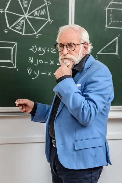 Nachdenkliche graue Haare Professor steht an der Tafel mit einem Stück Kreide — Stockfoto