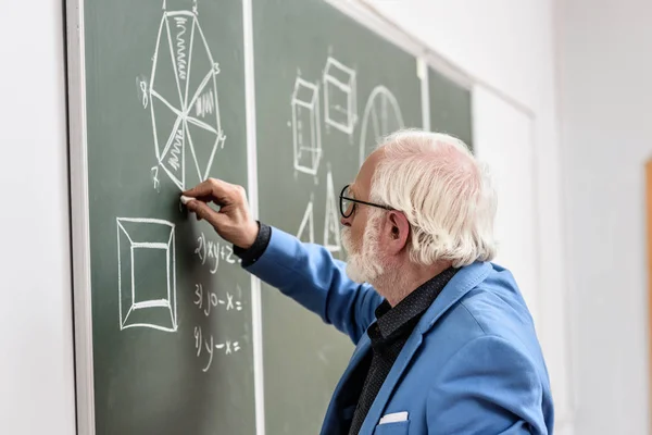 Vue latérale de professeur de cheveux gris dessin avec morceau de craie au tableau noir — Photo de stock