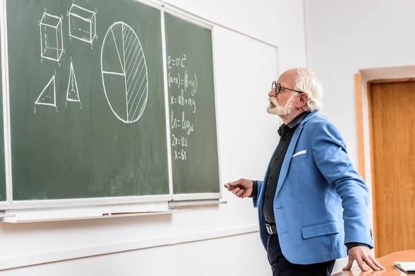 Grey hair professor holding piece of chalk and looking at blackboard — Stock Photo