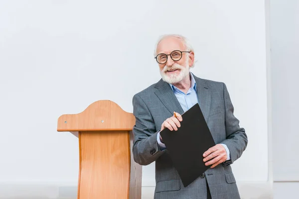 Sonriente profesor senior sosteniendo portapapeles y mirando a la cámara - foto de stock