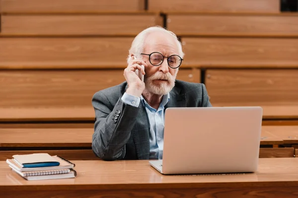 Professor de cabelo cinza falando por smartphone e olhando para laptop — Fotografia de Stock