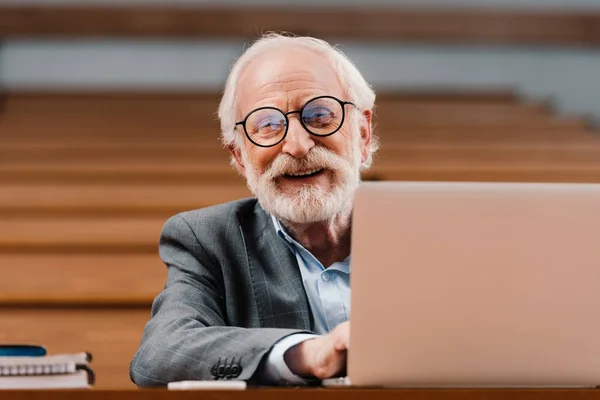 Sorridente cinza professor de cabelo sentado em sala de aula vazia com laptop — Fotografia de Stock